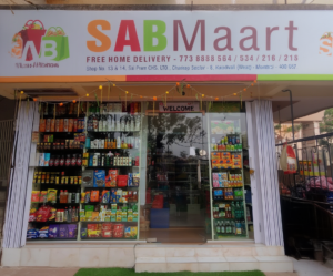 Front view of a small grocery store with a colorful sign reading "SABMaart." Shelves are filled with various packaged goods. A welcome sign and festive lights add a warm, inviting tone.