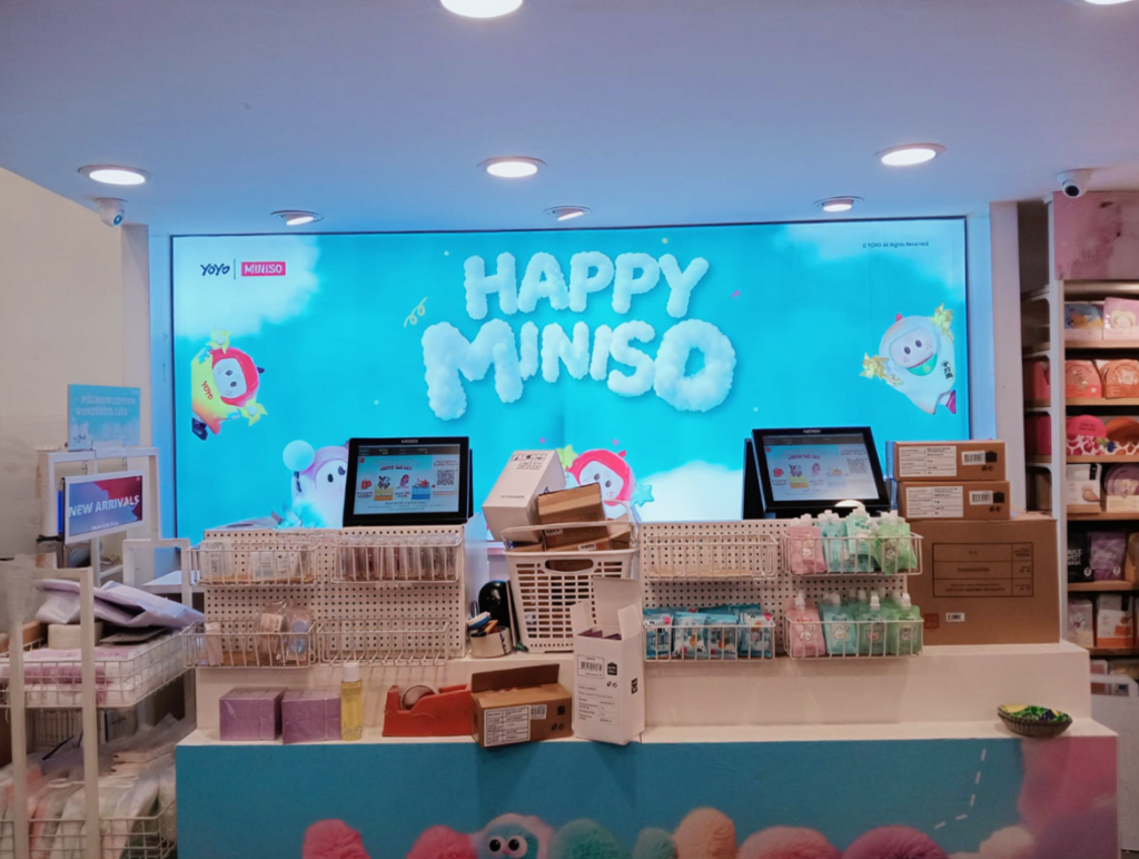 Toy store checkout counter with bright "Happy Miniso" sign. Baskets hold pastel items; colorful plush toys displayed, evoking a cheerful vibe.