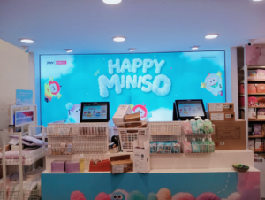 Toy store checkout counter with bright "Happy Miniso" sign. Baskets hold pastel items; colorful plush toys displayed, evoking a cheerful vibe.
