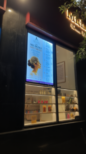 A warmly lit shop window displays beauty products neatly arranged on shelves. A large sign shows a profile of a woman with text about the "Michewa Method."